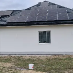 Biały dom jednorodzinny z zamontowanymi panelami słonecznymi na ciemnym dachu, widok z boku, z widocznym wiaderkiem farby u dołu kadru, sugerującym świeżo pomalowaną elewację.