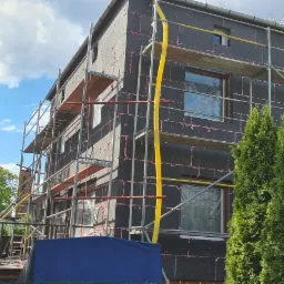 Dom w trakcie ocieplania, z widocznym styropianem na elewacji, rusztowaniami i zabezpieczonymi oknami, otoczony zielenią i błękitnym niebem.