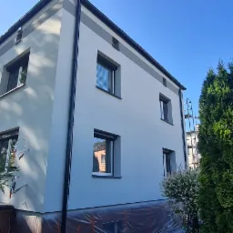 Jasny, dwupiętrowy dom z szarą stolarką okienną i wykończeniem dachu, otoczony zielenią, z widocznymi śladami prac elewacyjnych w postaci zabezpieczonej folią podstawy budynku i drabiny w tle.