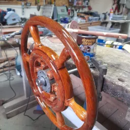 Lakierowane drewniane koło sterowe na warsztatowym stole, widoczna puszka z bejcą i narzędzia w tle.