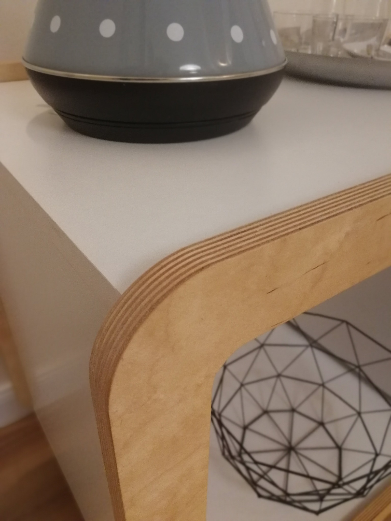 Fragment blatu z jasnego drewna warstwowego z zaokrąglonym rogiem, na którym stoi szary czajnik w białe kropki; w dolnej półce widoczny druciany kosz.