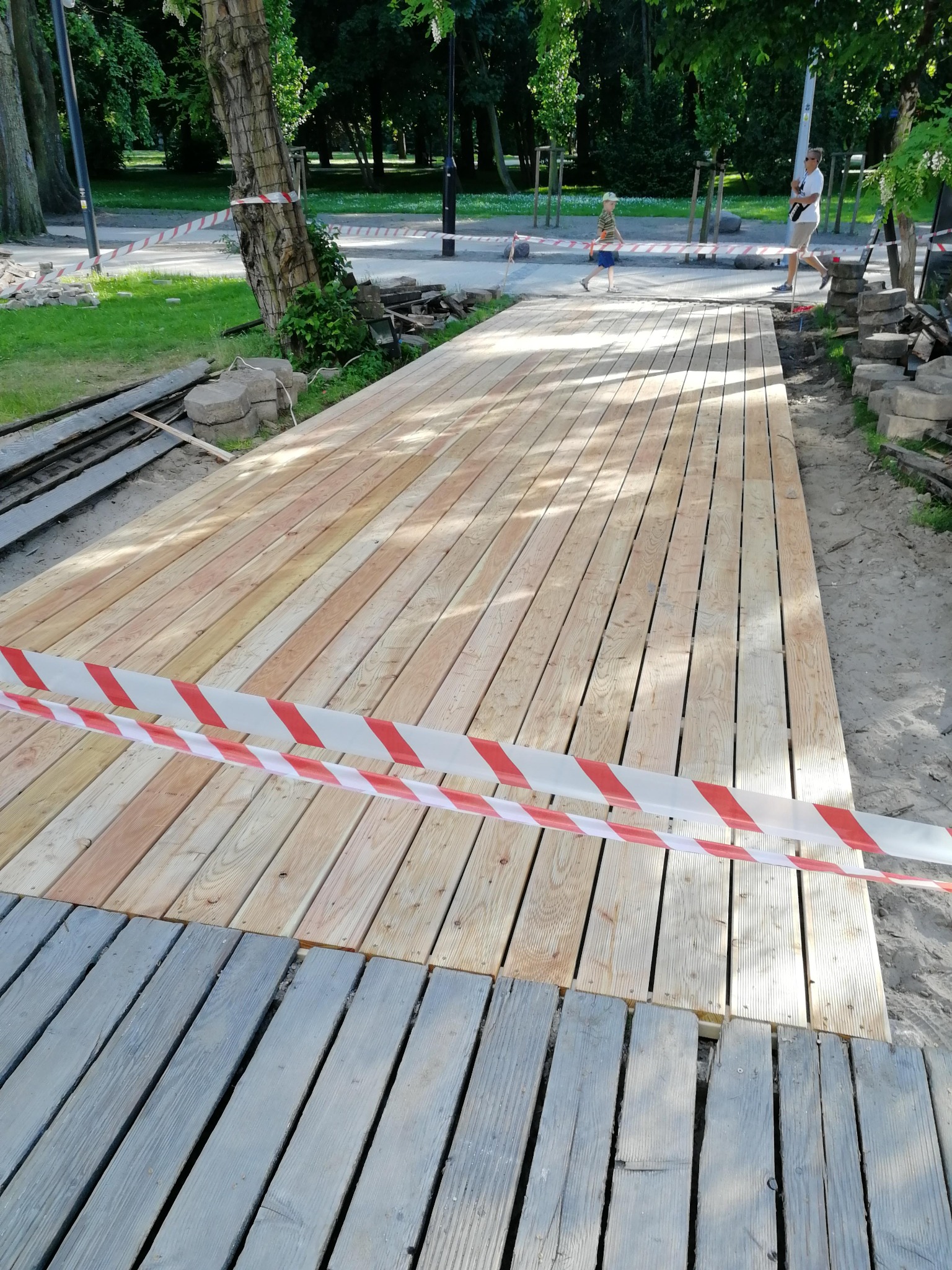 Nowa drewniana ścieżka w parku w trakcie budowy, otoczona taśmą ostrzegawczą, z widocznymi elementami starej, szarej ścieżki i przygotowanym podłożem.