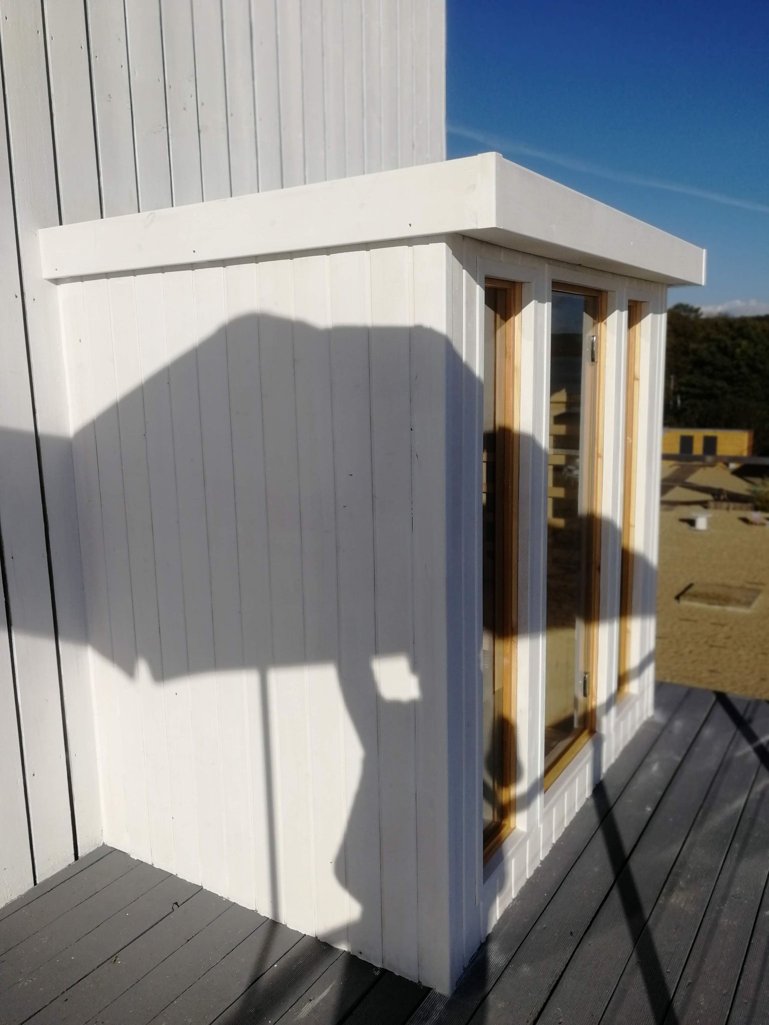 Biała, drewniana zabudowa okienna z dwoma oknami, widoczna z zewnątrz na tle pomalowanej na szaro drewnianej podłogi i niebieskiego nieba, rzucająca cień na ścianę z desek.