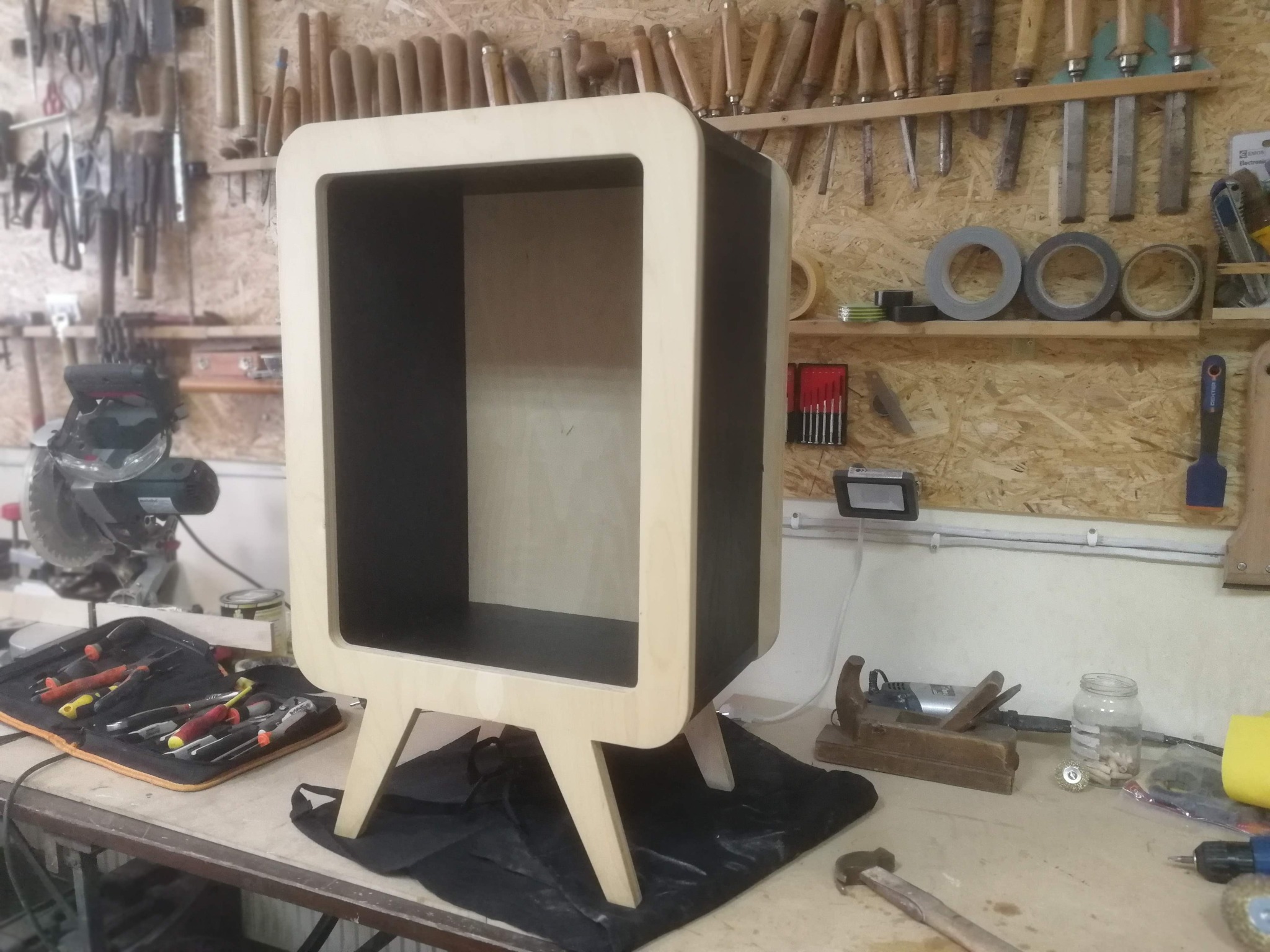 Niewykończona szafka nocna z jasnego drewna z zaokrąglonymi krawędziami i czarnym wnętrzem, stojąca na warsztatowym stole obok narzędzi.