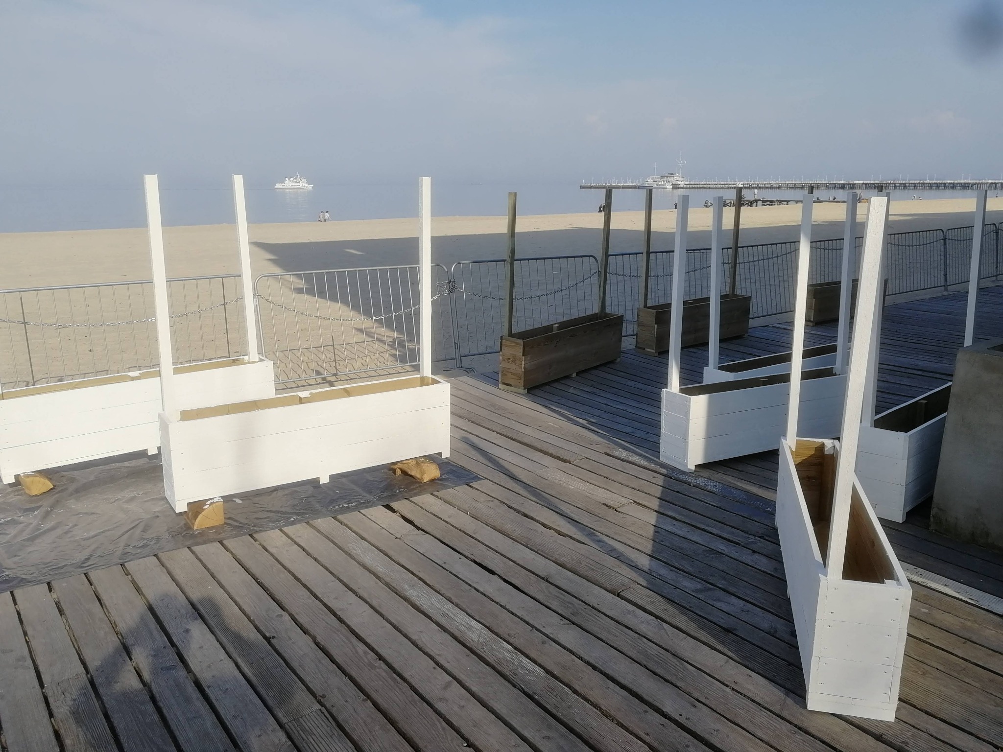 Białe, drewniane donice z pionowymi słupkami, ustawione na drewnianym pomoście, z widokiem na plażę i morze w tle.