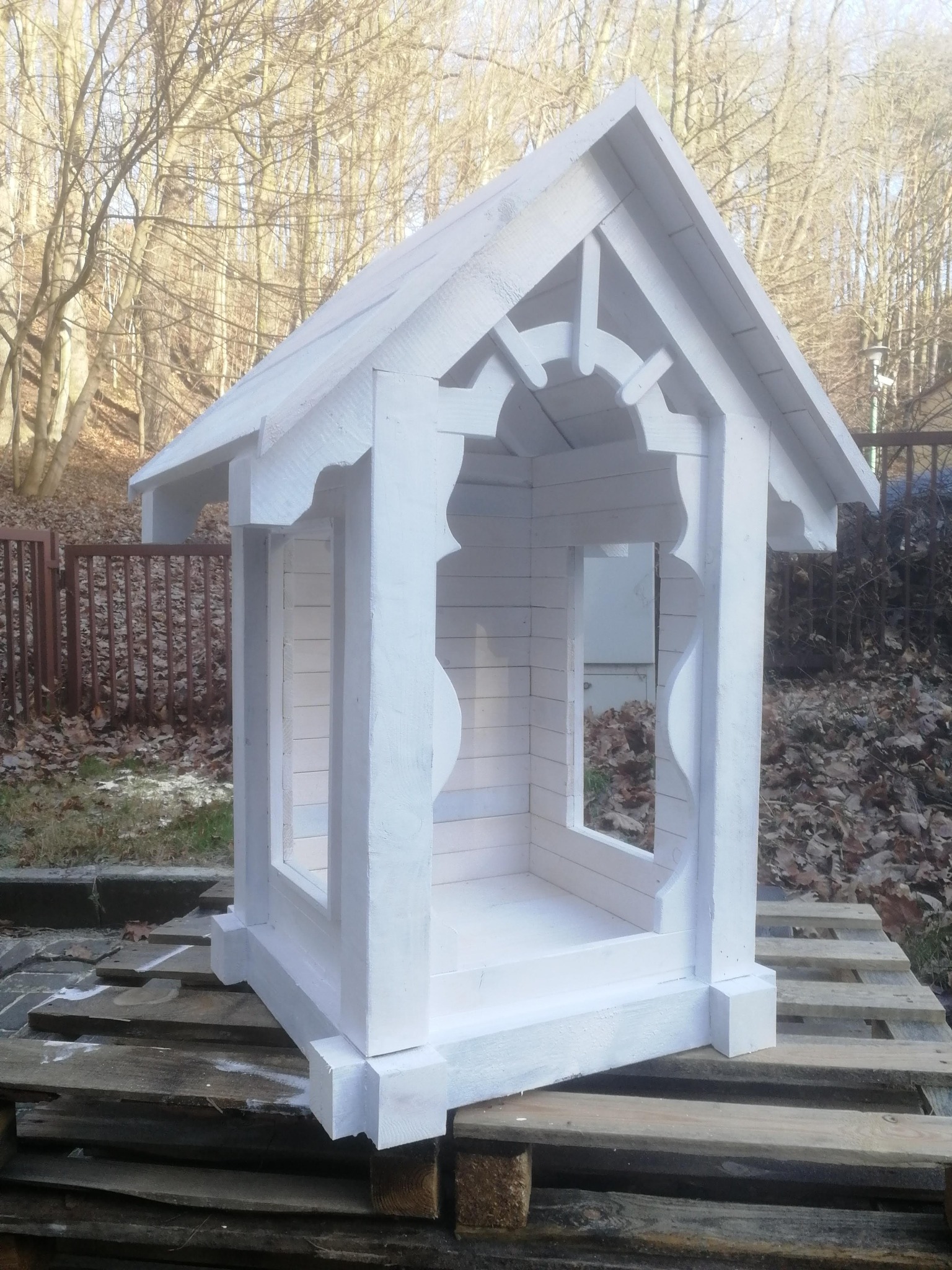 Biała, drewniana konstrukcja przypominająca domek dla lalek lub karmnik dla ptaków, z dekoracyjnymi wycięciami, ustawiona na palecie na tle drzew bez liści.