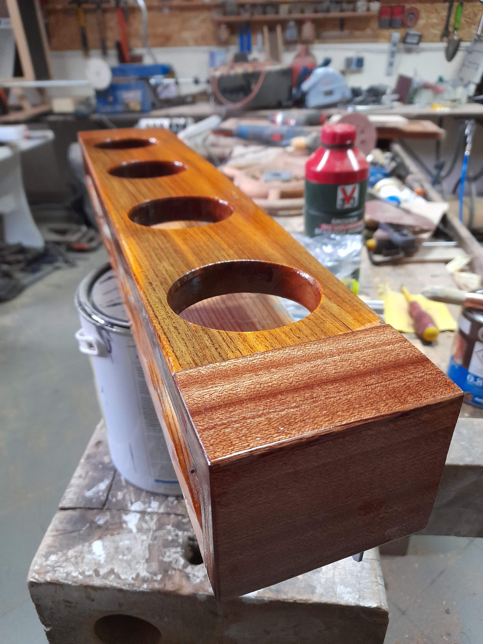 Lakierowany drewniany element z czterema otworami, prawdopodobnie stojak na coś, leży na warsztatowym stole w tle widoczne narzędzia i materiały.