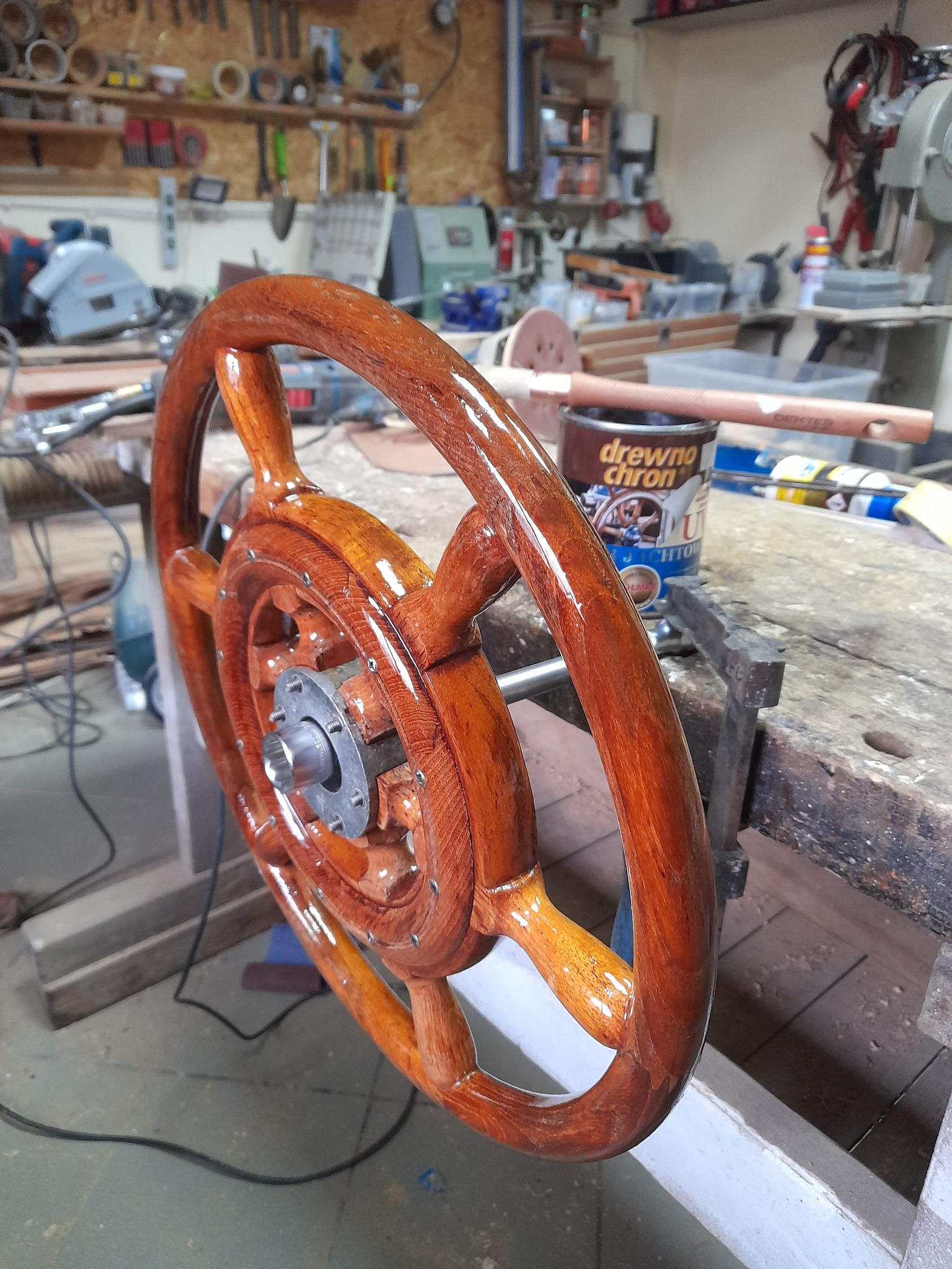 Lakierowane drewniane koło sterowe na warsztatowym stole, widoczna puszka z bejcą i narzędzia w tle.