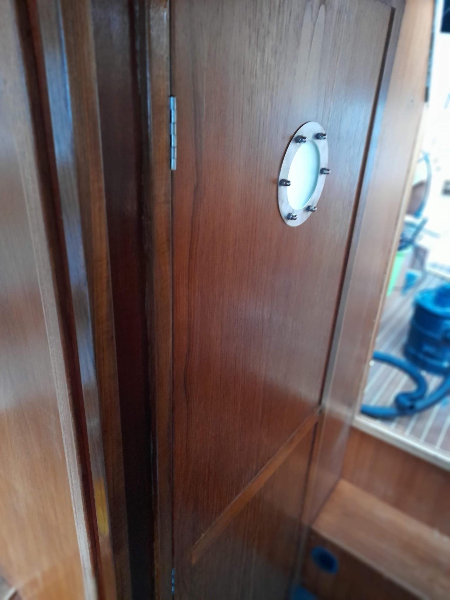 Drewniane drzwi z okrągłym okienkiem z metalową ramką, zamontowane w drewnianej zabudowie jachtu, widoczne zawiasy i fragment drewnianego pokładu.