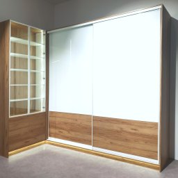 WROMAX Maciej Wroński - Nowoczesna szafa narożna z przesuwnymi, białymi drzwiami i drewnianymi akcentami, podświetlana od dołu, z przeszkloną witryną z półkami po lewej stronie.