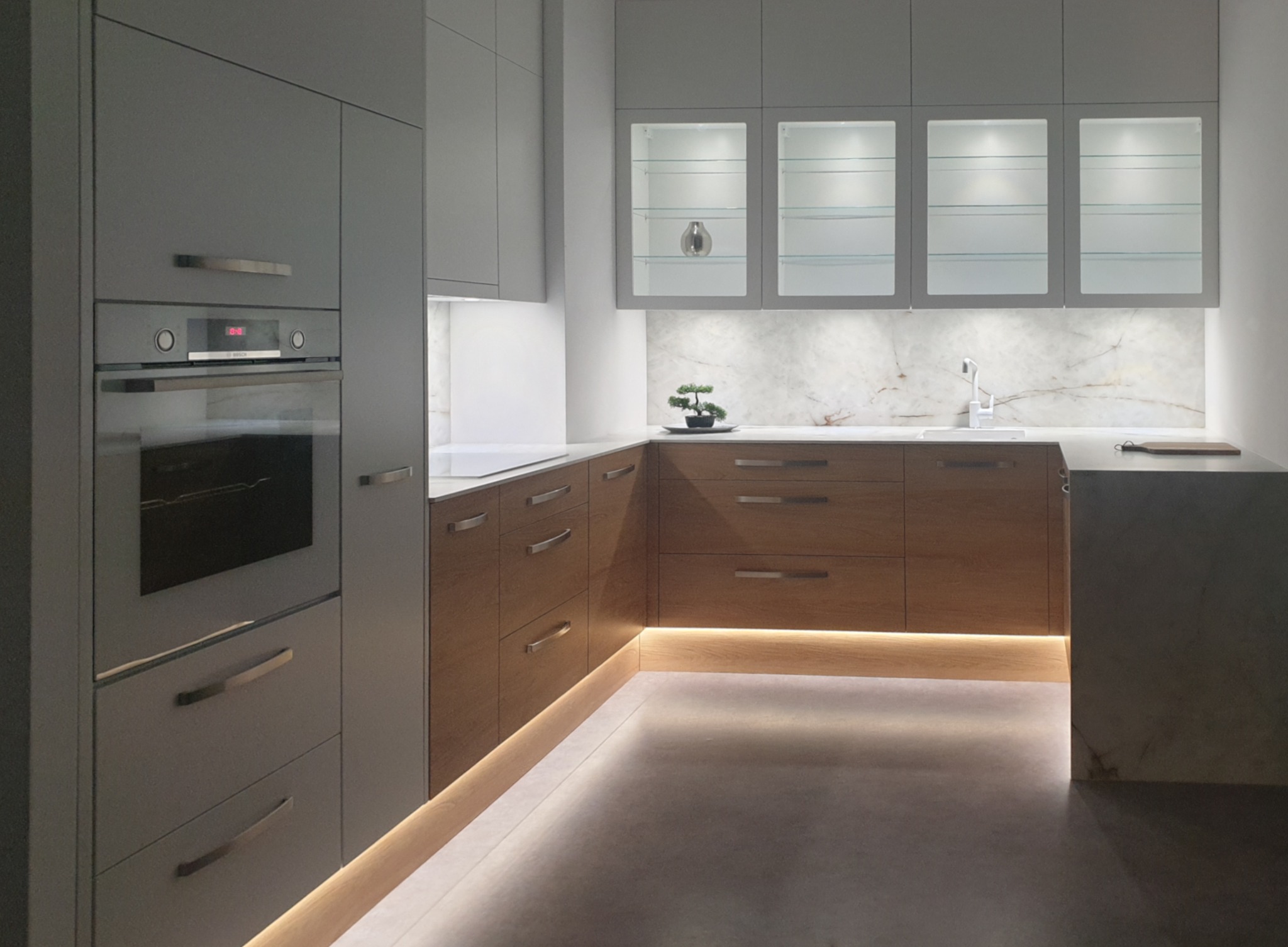 Nowoczesna kuchnia z szarymi szafkami i drewnianymi frontami, podświetleniem LED przy podłodze i blacie, marmurowym backsplashiem i szklanymi witrynami.