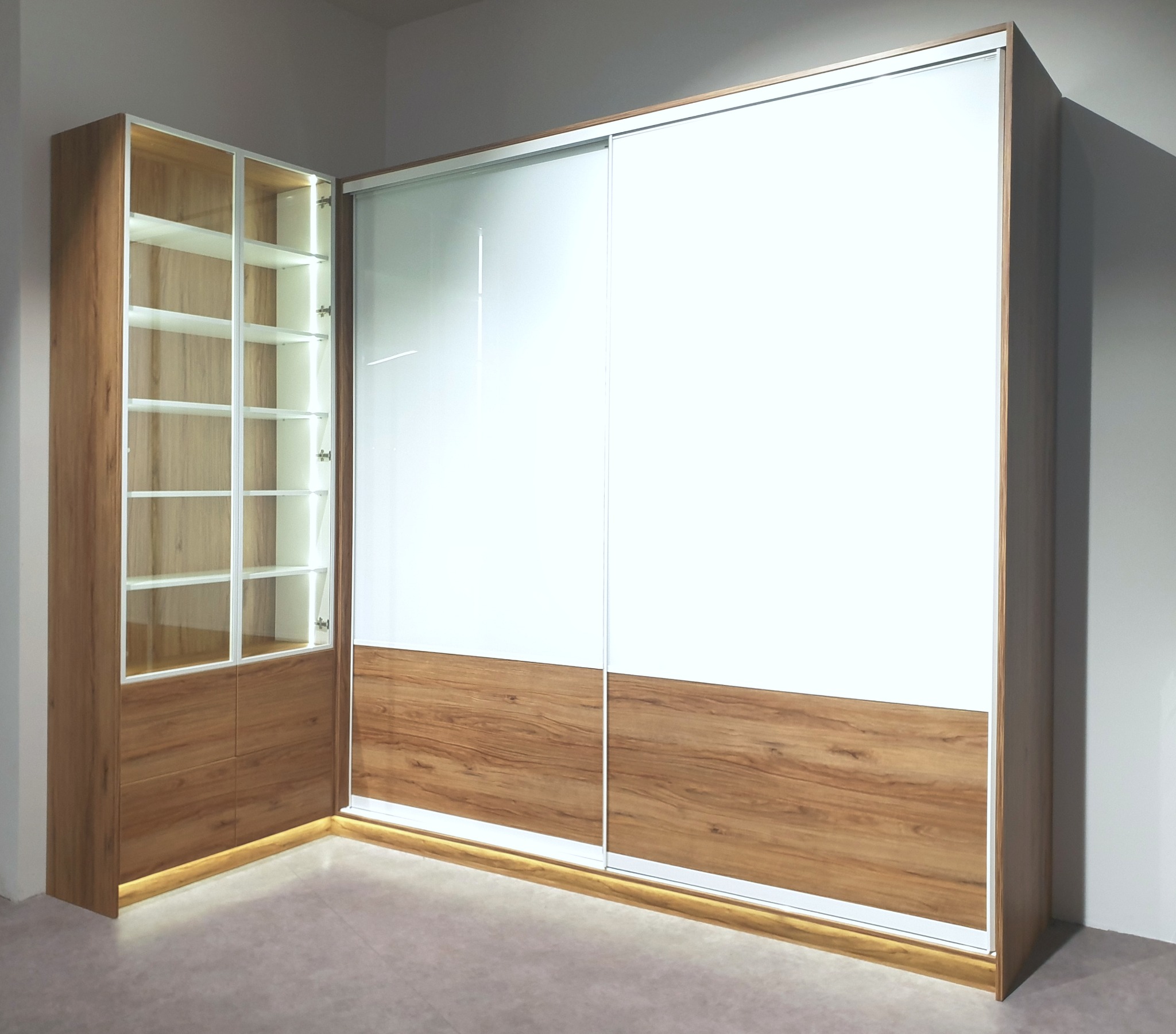 Nowoczesna szafa narożna z przesuwnymi, białymi drzwiami i drewnianymi akcentami, podświetlana od dołu, z przeszkloną witryną z półkami po lewej stronie.