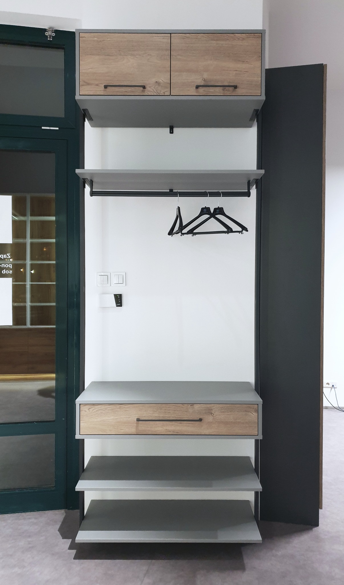 Wnęka wejściowa z szafkami z drewnopodobnym frontem, szarą półką i szufladą, wieszakiem na ubrania i dwiema szarymi półkami na buty, obok drzwi wejściowych.