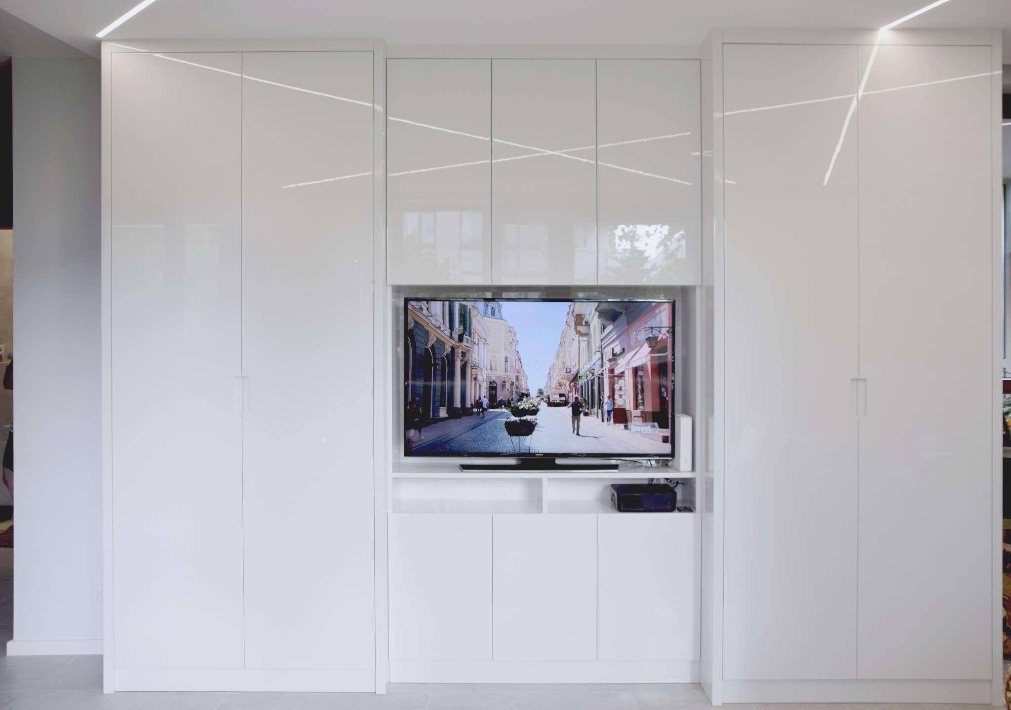 Biała zabudowa meblowa z szafami po bokach i wnęką na telewizor prezentujący widok ulicy, minimalistyczny design z podświetleniem LED.