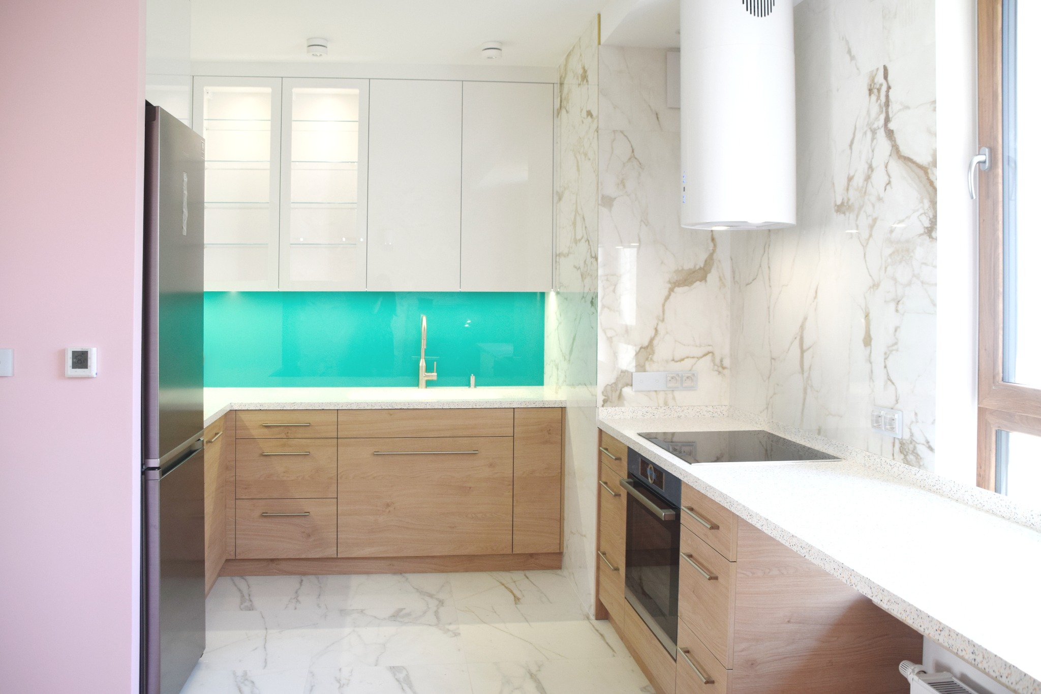 Nowoczesna kuchnia z drewnianymi szafkami, białymi górnymi szafkami, turkusowym panelem szklanym i marmurowym wykończeniem ściany.