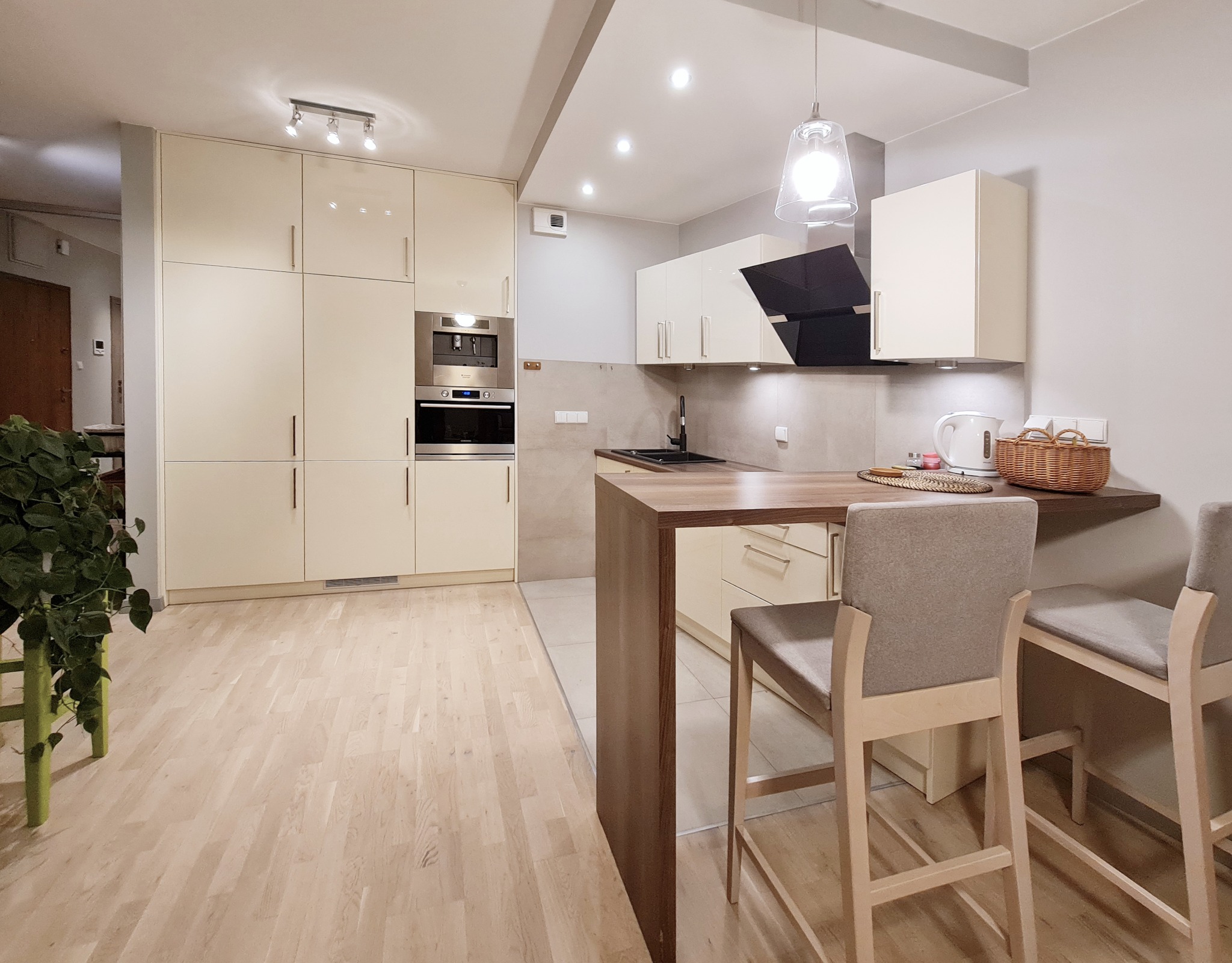 Jasna kuchnia z zabudową na wymiar, wyspą z drewnianym blatem, krzesłami barowymi i oświetleniem punktowym.