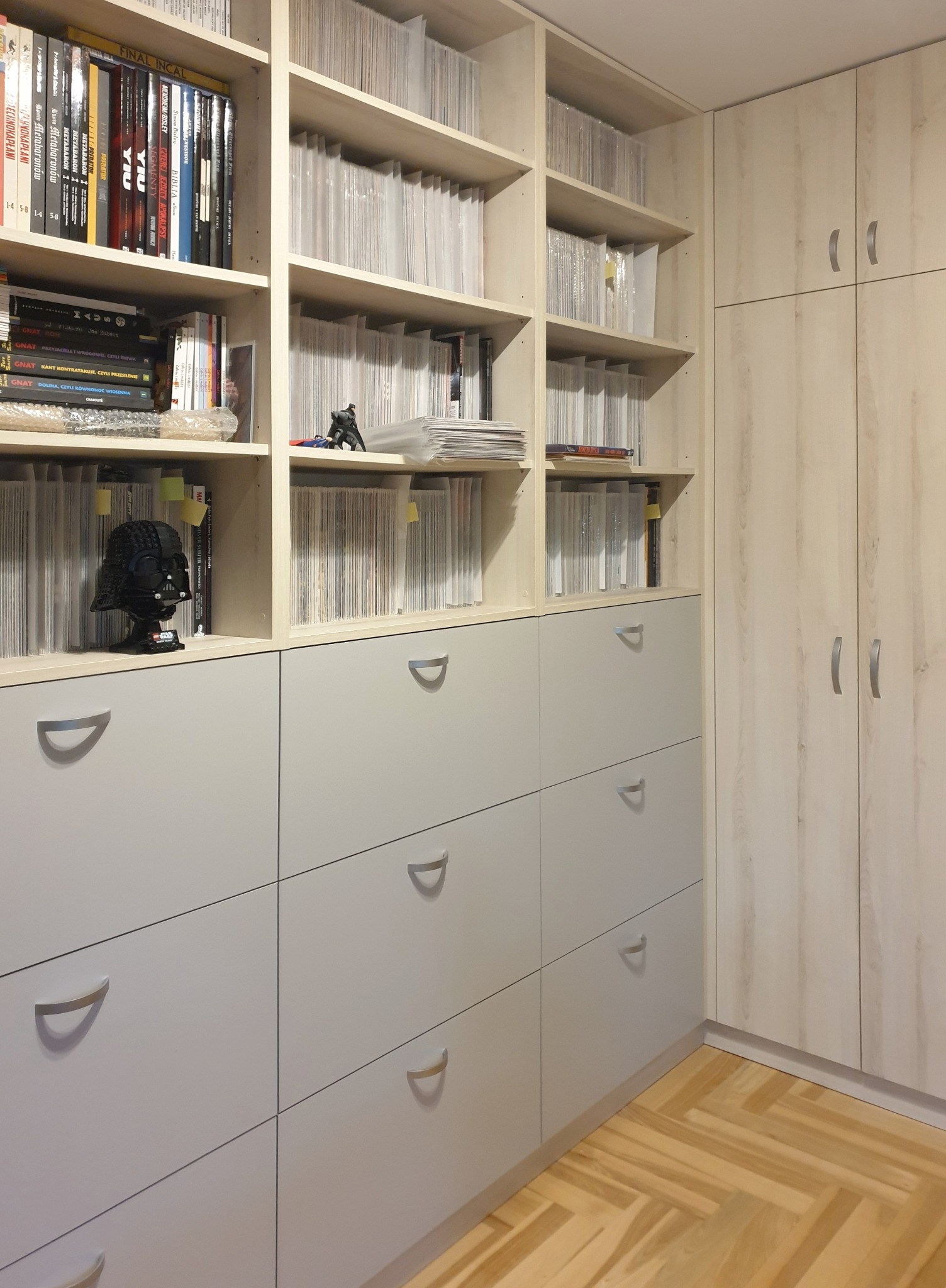 Regał z szufladami i półkami wypełnionymi książkami i komiksami, z figurką Dartha Vadera z klocków Lego na jednej z półek. Obok znajduje się szafa z jasnego drewna, a na podłodze parkiet ułożony...