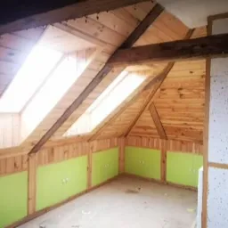 Poddasze wykończone boazerią i drewnianymi belkami, z oknami dachowymi i zielonymi szafkami wbudowanymi w ścianę, fragment ściany w surowym stanie przed wykończeniem.