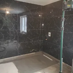 Łazienka w trakcie remontu: czarne, marmurowe płytki ścienne z białymi żyłami, wylewka podłogowa, poziomica laserowa, okno w tle.