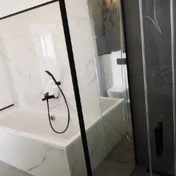 Nowoczesna łazienka z wanną obudowaną białymi płytkami imitującymi marmur, czarna bateria wannowa z prysznicem oraz kabina prysznicowa z ciemnymi płytkami i czarnymi profilami.