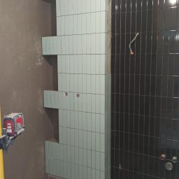 Dmytro Vlasov Sova VDV - Niebiesko-czarna łazienka w trakcie wykańczania. Widoczne ułożone płytki z wnękami, poziomica laserowa, szlifierka kątowa i krzyżyki dystansowe.