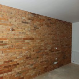 Dmytro Vlasov Sova VDV - Ściana wykończona czerwoną cegłą klinkierową w surowym wnętrzu. Widoczne gniazdka elektryczne i niedokończone podłoże. Oświetlenie sufitowe w tle.