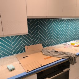 Dmytro Vlasov Sova VDV - Turkusowa glazura ułożona w jodełkę na ścianie kuchni, widoczne narzędzia i karton OBI na blacie, białe szafki kuchenne. Etap prac wykończeniowych.