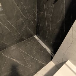 Dmytro Vlasov Sova VDV - Wykończona ściana w łazience z czarnych płytek imitujących marmur, widoczne przyłącza wodne i fragment białej umywalki. Minimalistyczny design.