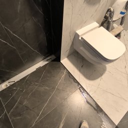 Dmytro Vlasov Sova VDV - Wykończona łazienka z czarno-białymi płytkami imitującymi marmur, wisząca toaleta. Widoczny odpływ liniowy i fragment drabiny malarskiej.