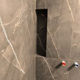 Dmytro Vlasov Sova VDV - Wykończona ściana w łazience z szarych płytek imitujących marmur z czarną wnęką i wystającymi rurami z ciepłą i zimną wodą. Minimalistyczny design.