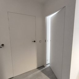 Dmytro Vlasov Sova VDV - Minimalistyczne wnętrze: białe, ukryte drzwi z czarną klamką, szara podłoga, sufit z oświetleniem punktowym. Jedne drzwi otwarte, ukazują kolejne pomieszczenie.