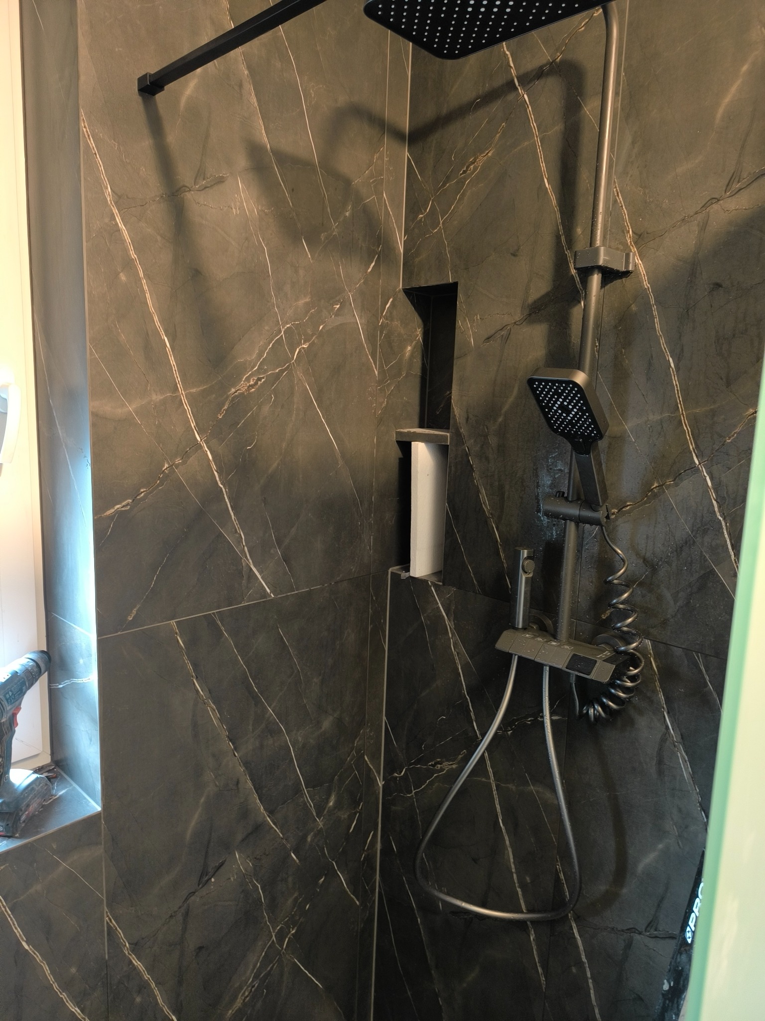 Nowoczesna kabina prysznicowa z czarną armaturą i płytkami imitującymi marmur. Widoczny fragment wiertarki sugeruje prace wykończeniowe.