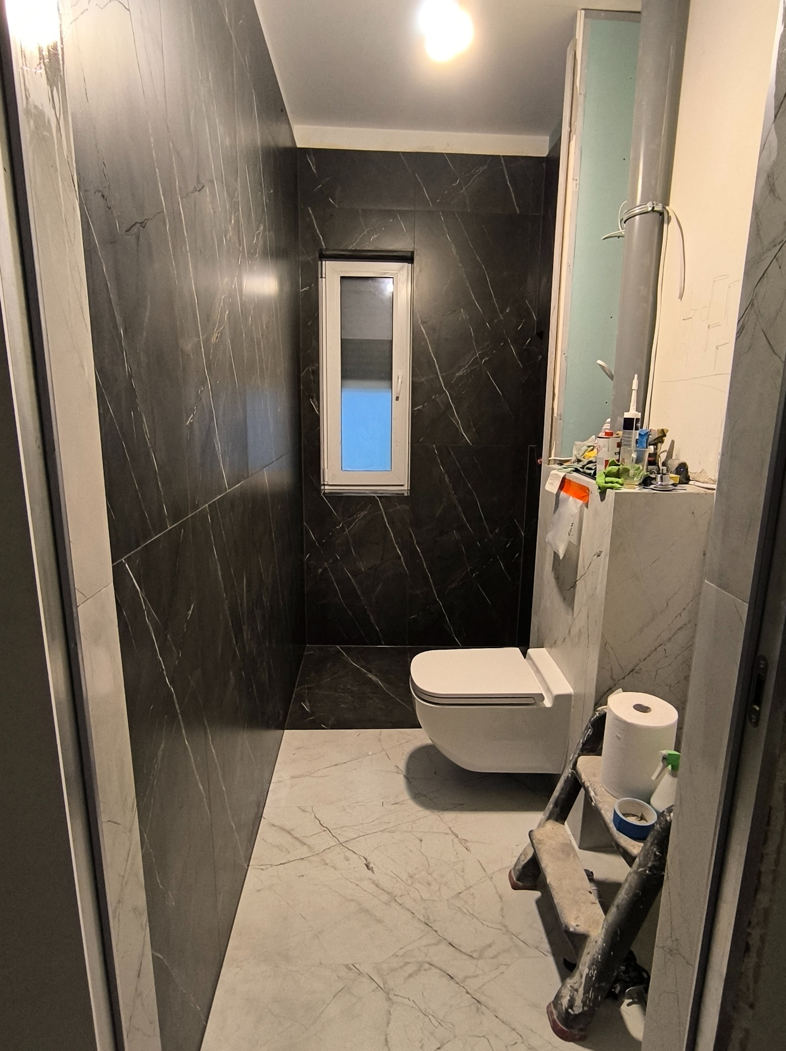 Remont łazienki w trakcie: czarne płytki imitujące marmur na ścianach, biała wisząca toaleta, drabina malarska i akcesoria. Widok na okno i fragment instalacji.