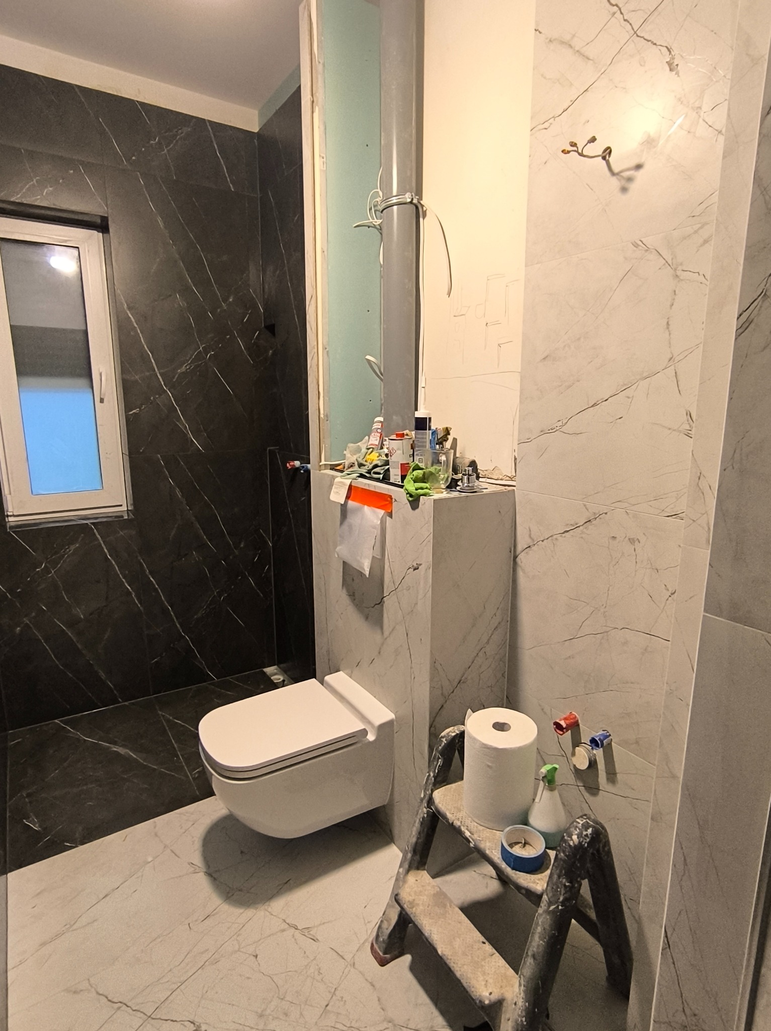 Remont łazienki w trakcie: biała wisząca toaleta na tle marmurowych płytek, drabina z akcesoriami, widoczne rury i niedokończone ściany. Ujęcie z bliska.