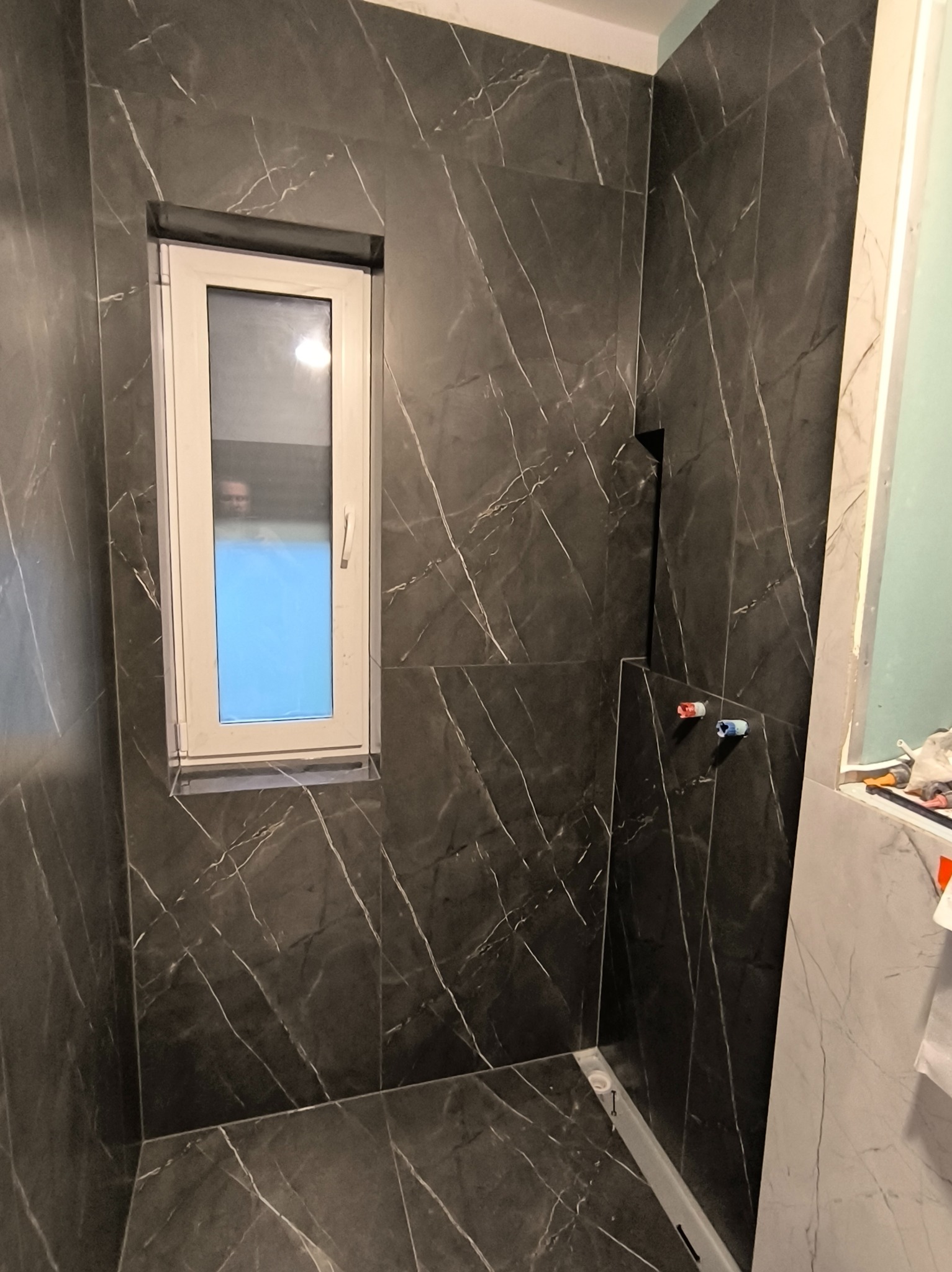 Wykończona łazienka z oknem, ściany i podłoga wyłożone ciemnymi płytkami imitującymi marmur z jasnymi żyłami. Widoczne elementy instalacji wodnej i odpływ liniowy.