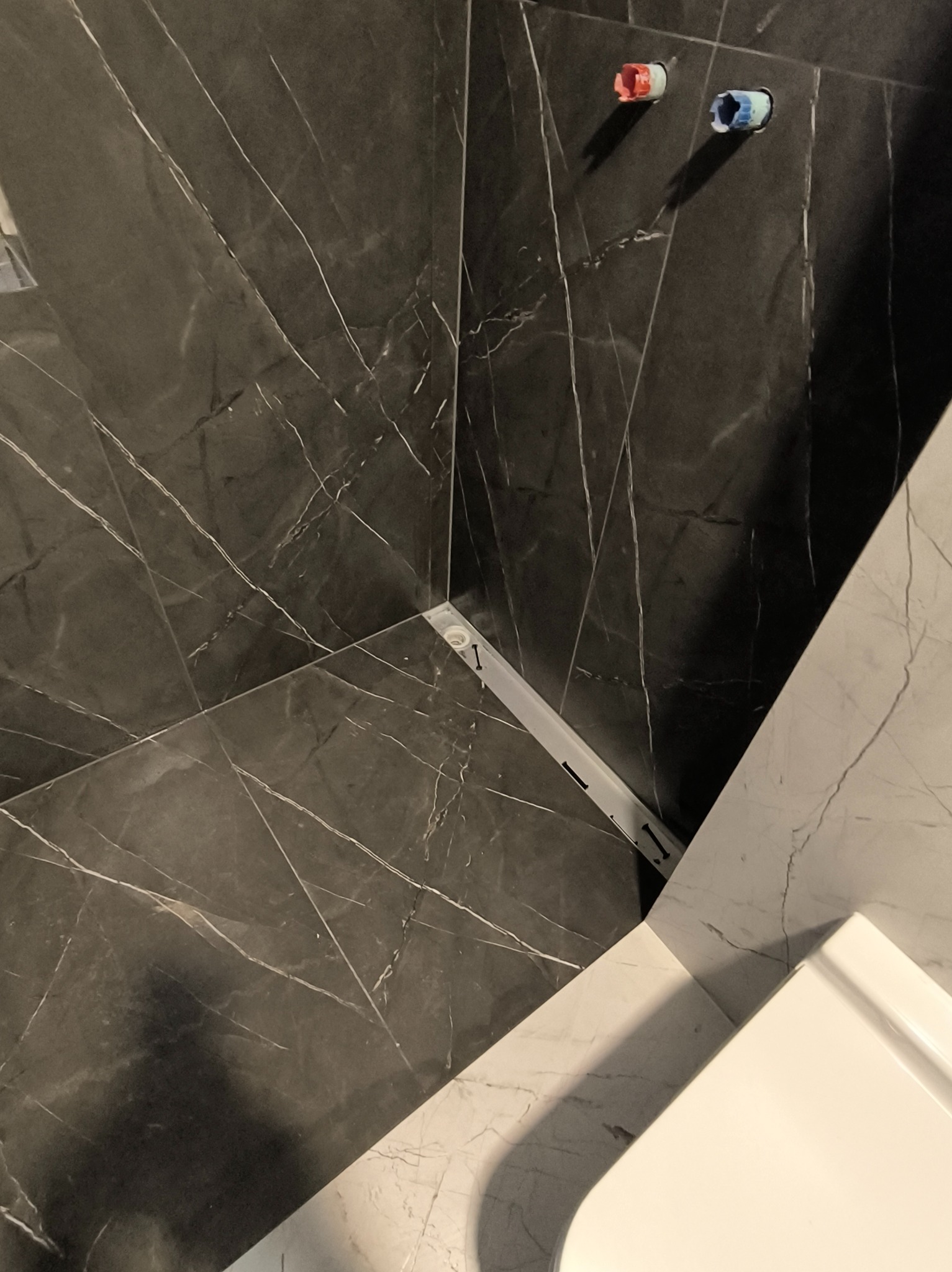 Wykończona ściana w łazience z czarnych płytek imitujących marmur, widoczne przyłącza wodne i fragment białej umywalki. Minimalistyczny design.