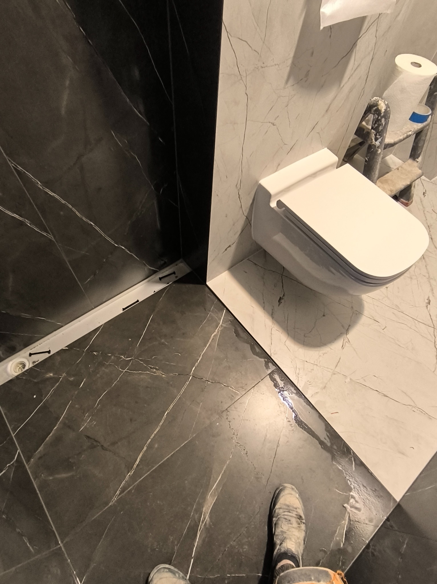 Wykończona łazienka z czarno-białymi płytkami imitującymi marmur, wisząca toaleta. Widoczny odpływ liniowy i fragment drabiny malarskiej.