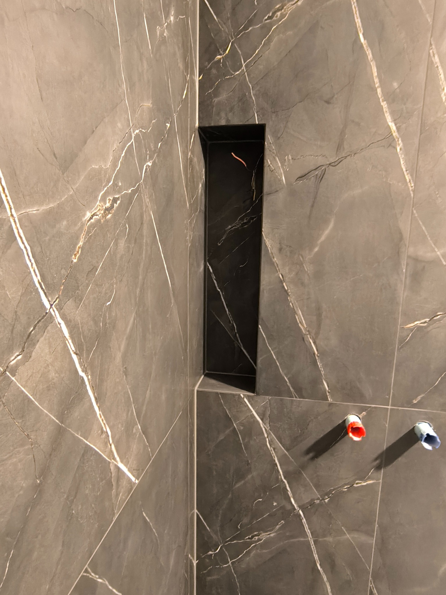 Wykończona ściana w łazience z szarych płytek imitujących marmur z czarną wnęką i wystającymi rurami z ciepłą i zimną wodą. Minimalistyczny design.