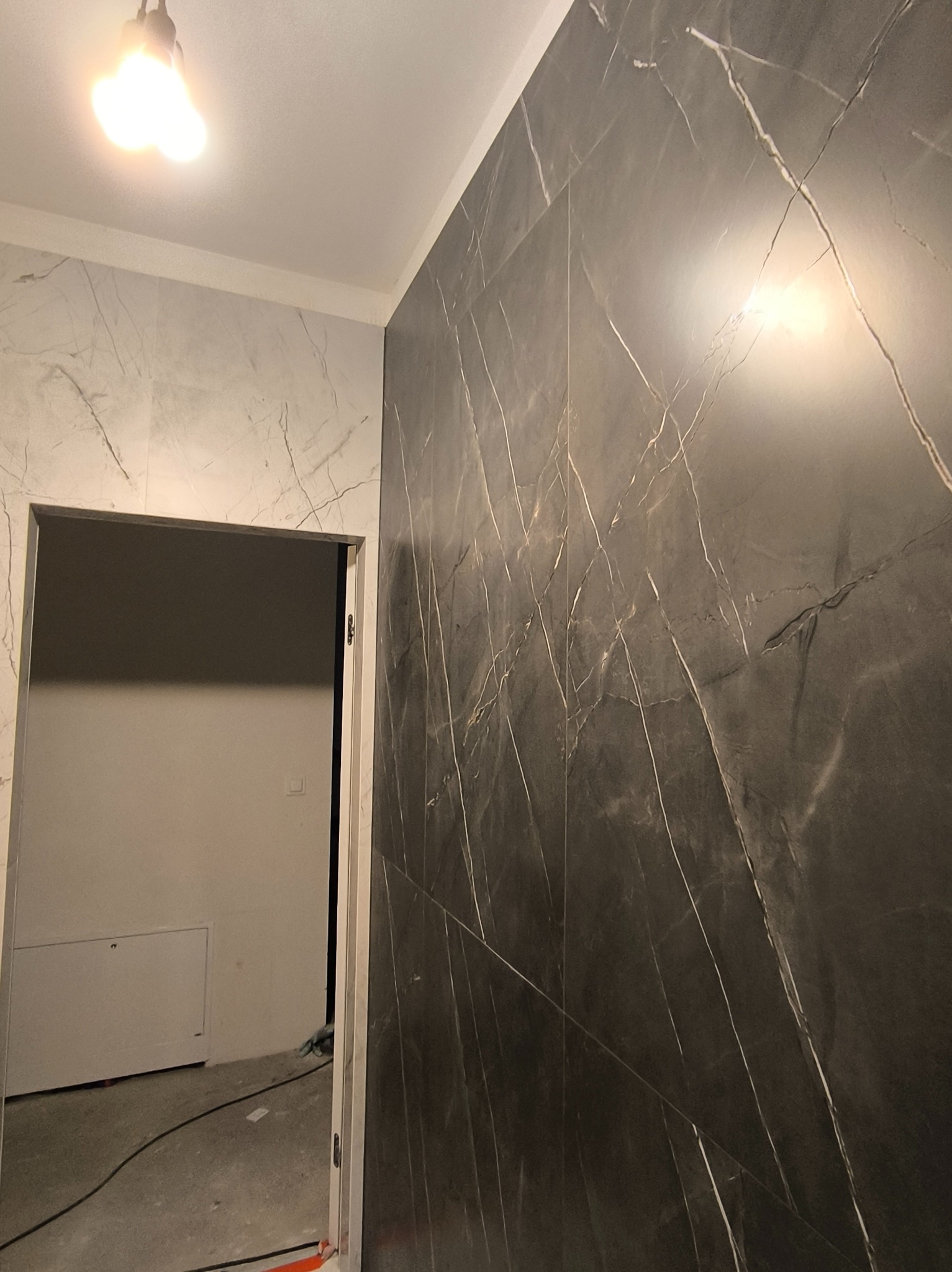 Ciemna, marmurowa okładzina ścienna z białymi żyłami w łazience, widok na otwarte drzwi i fragment ściany z jasnymi płytkami imitującymi marmur.