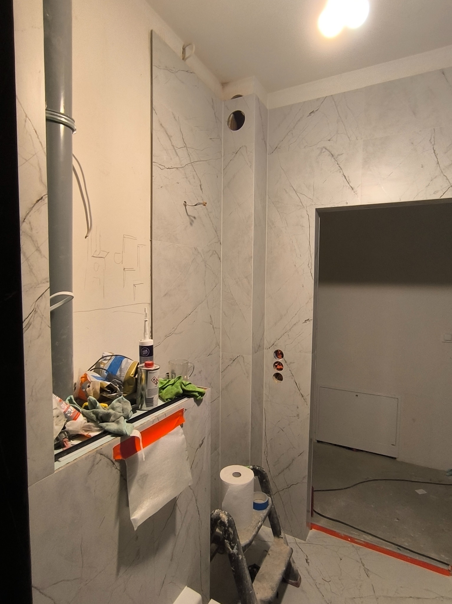 Łazienka w trakcie remontu: ściany wyłożone płytkami imitującymi marmur, widoczne narzędzia i materiały budowlane, drabina oraz instalacja elektryczna w ścianie.