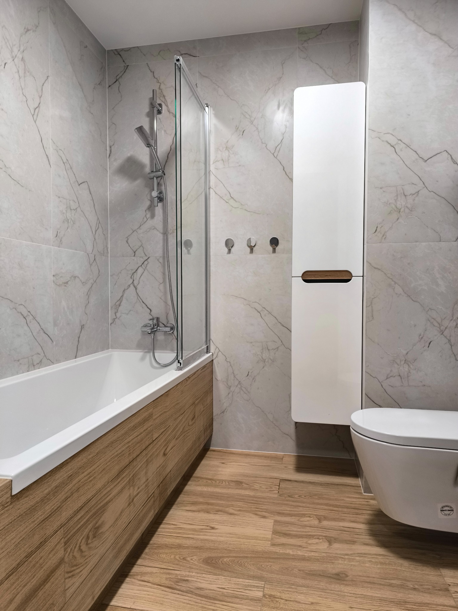 Nowoczesna łazienka z wanną obudowaną drewnem, szklanym parawanem, marmurowymi płytkami i podwieszaną szafką. Uwagę zwraca drewniana podłoga i minimalistyczne wieszaki.