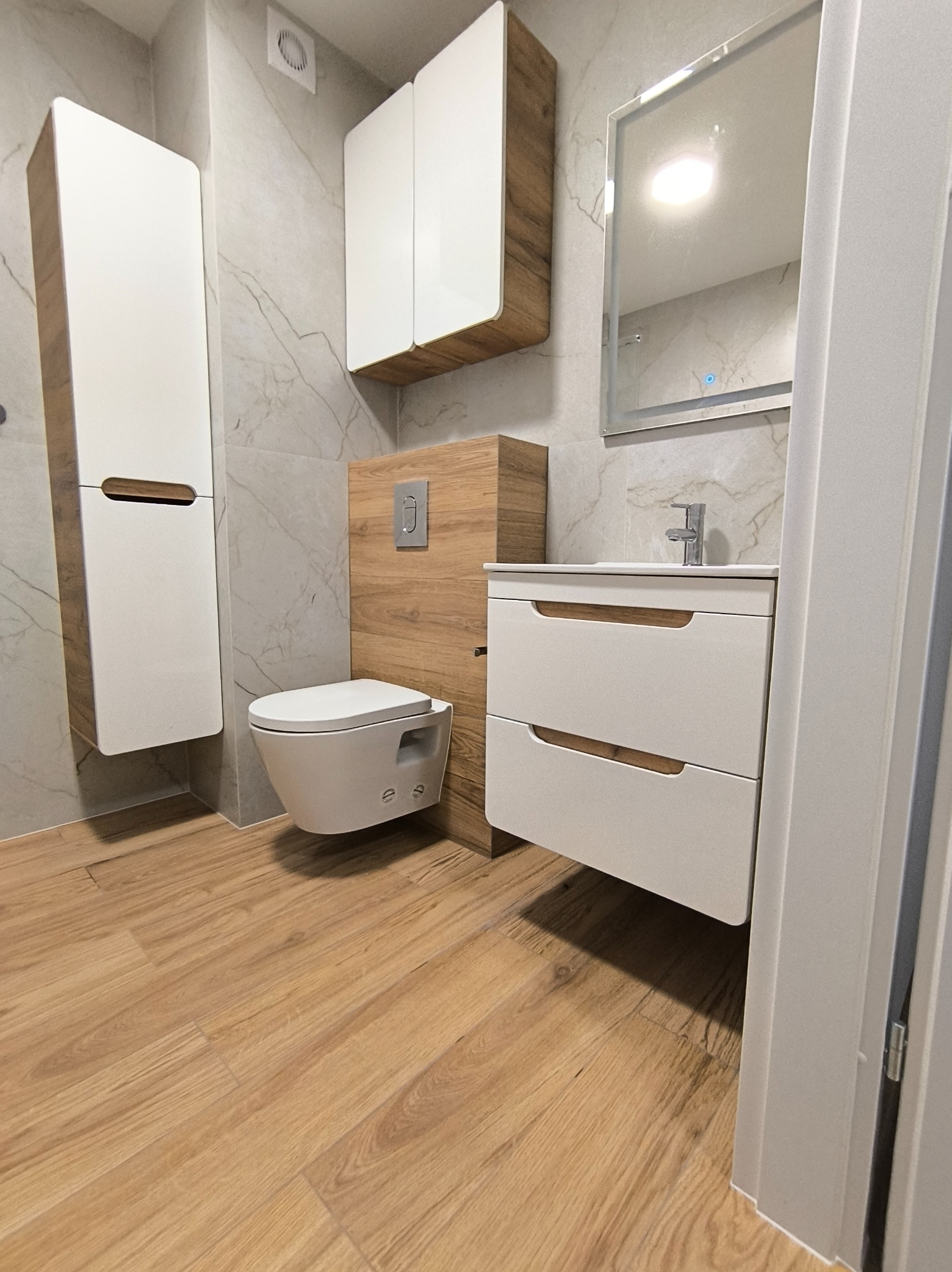 Nowoczesna łazienka po remoncie: podwieszana toaleta, szafka z umywalką, lustro z podświetleniem LED i drewnopodobna podłoga. Minimalistyczny design.