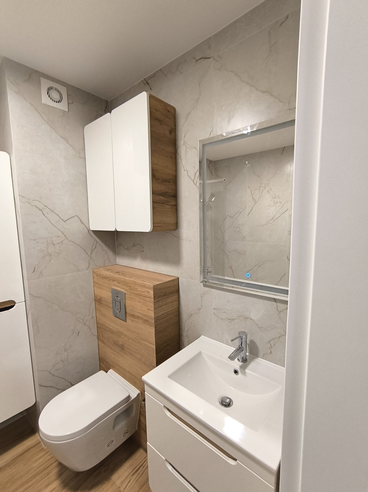 Nowoczesna łazienka z marmurowymi płytkami, wiszącą toaletą i szafkami z drewnianymi akcentami. Lustro z podświetleniem LED. Minimalistyczny design.