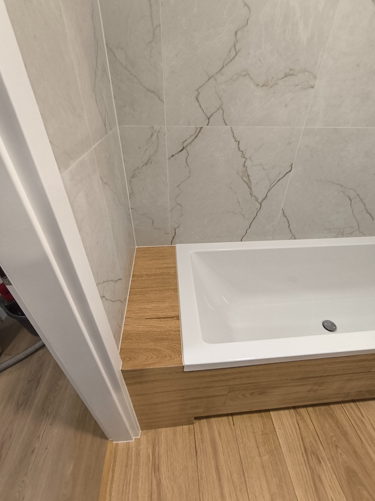 Nowoczesna łazienka: biała wanna z drewnianą obudową, marmurowe płytki na ścianie i drewnopodobna podłoga. Minimalistyczny design, jasne kolory, świeżo po remoncie.