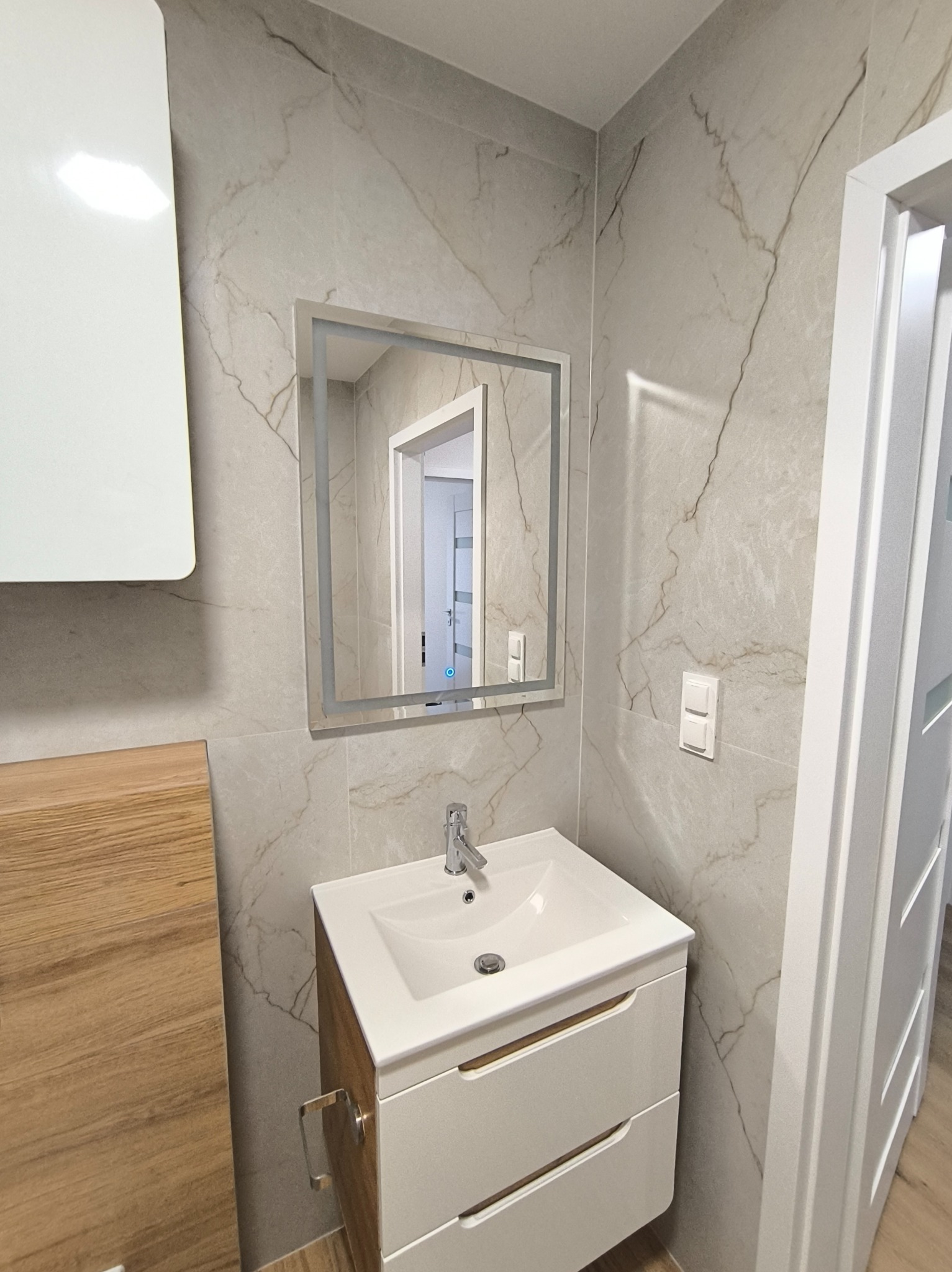 Wykończona łazienka z umywalką na szafce, lustrem z podświetleniem LED i płytkami imitującymi marmur. Widoczny fragment drzwi i szafki wiszącej.