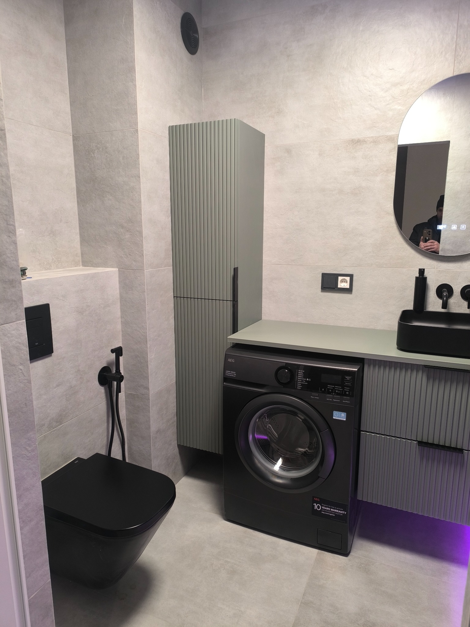 Nowoczesna łazienka w szarościach z czarną armaturą i pralką AEG. Umywalka nablatowa, podświetlenie LED i ryflowane fronty szafek dodają elegancji.