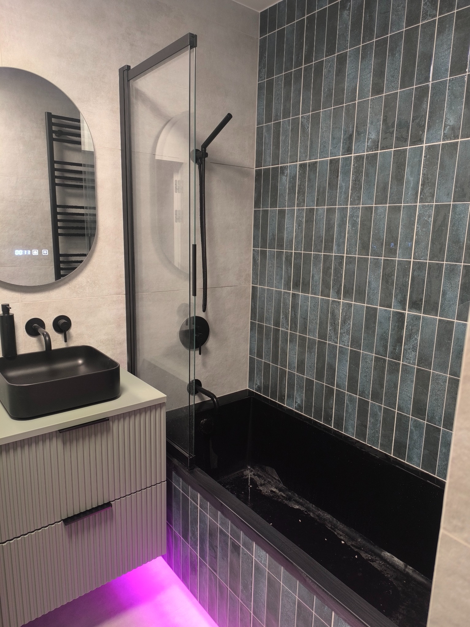 Nowoczesna łazienka z czarną armaturą, umywalką i wanną. Ściany wyłożone pionowymi, grafitowymi płytkami. Podświetlenie LED pod szafką umywalkową.