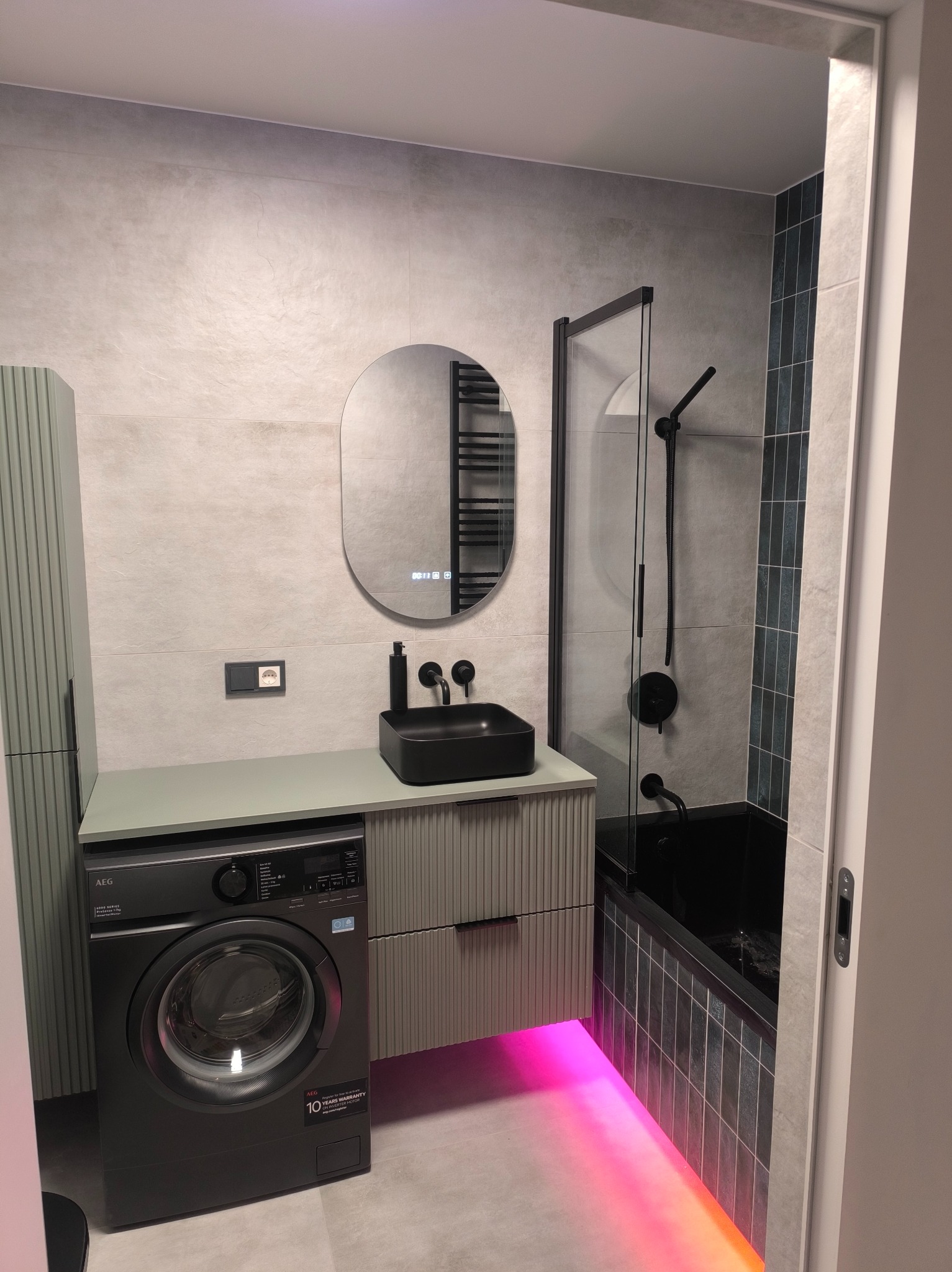 Nowoczesna łazienka z czarną armaturą, podświetleniem LED i pralką pod umywalką. Minimalistyczny design z szarymi płytkami i owalnym lustrem.