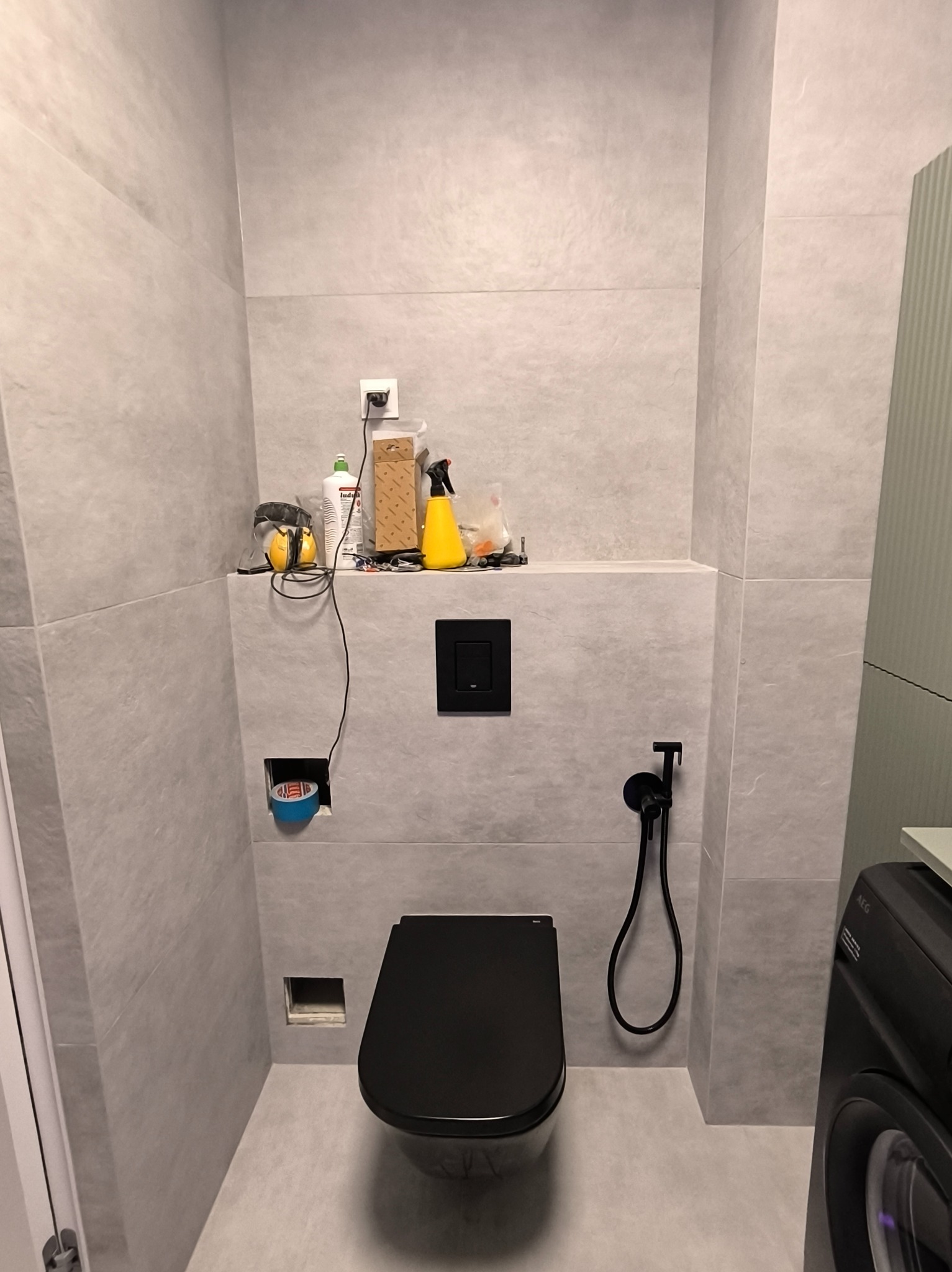 Szara łazienka z czarną toaletą podtynkową i bidetką. Na półce nad toaletą akcesoria i narzędzia. Widoczne niedokończone prace przy instalacji elektrycznej.
