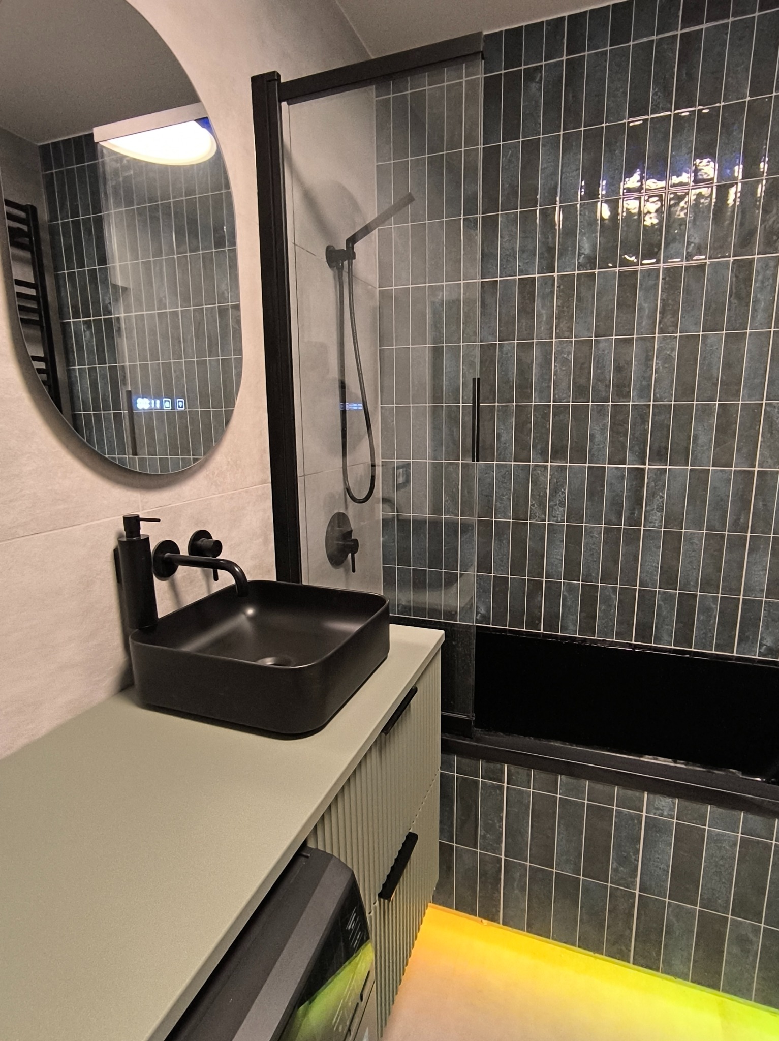 Nowoczesna łazienka z czarną umywalką nablatową, podświetlanym lustrem i kabiną prysznicową z ciemnymi płytkami ułożonymi pionowo.